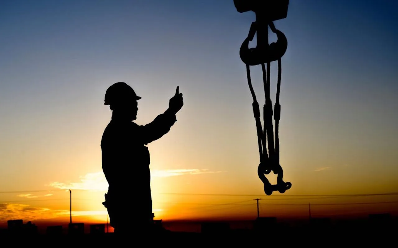 Crane crew member signalling beside suspended lifting gear at sunset